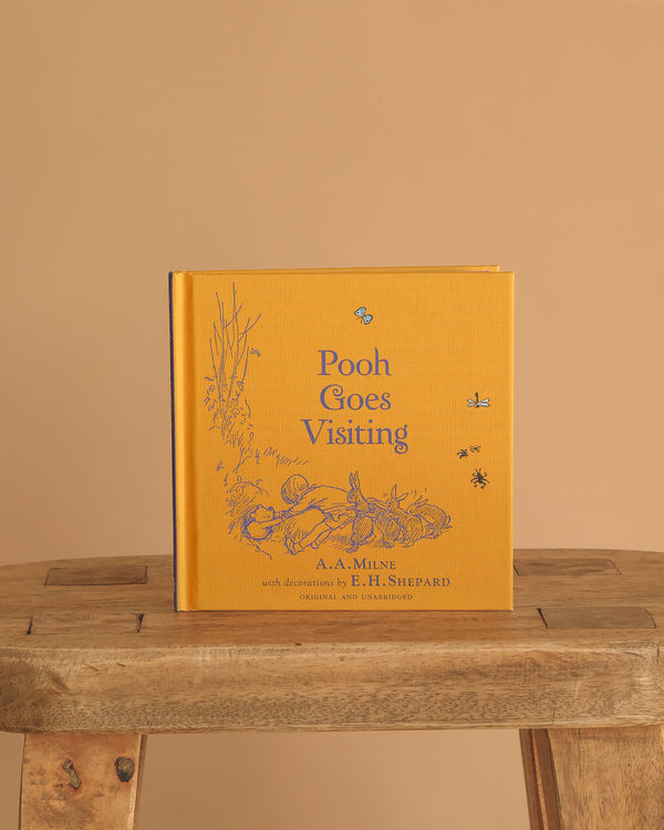 The yellow hardcover "Winnie-the-Pooh: Pooh Goes Visiting" stands upright on a rustic wooden stool against a beige background, featuring blue illustrations of Winnie-the-Pooh and friends on the cover.