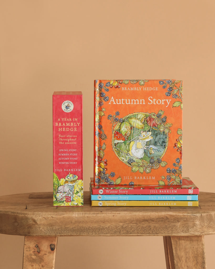 A stack of "A Year In Brambly Hedge" children's books by Jill Barklem sits on a wooden table, with one book displayed upright and the collection’s decorative slipcase shown against a beige background.