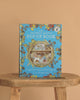 The Brambly Hedge Pop-Up Book by Jill Barklem stands upright on a wooden table against a beige background, its cover featuring vibrant illustrations of a mouse home and foliage on a blue backdrop.
