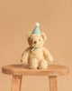 Snuggle Bunnies - Birthday Arthur - Sky is a plush beige teddy bear with a light blue party hat and ribbon, seated on a wooden stool against a tan background—a charming keepsake gift for birthdays.