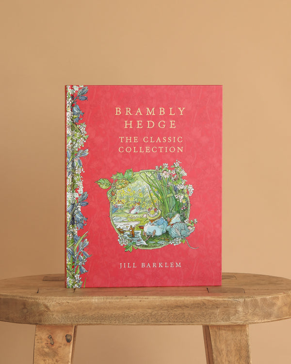 Brambly Hedge: The Classic Collection by Jill Barklem" is a red hardcover book with floral and nature illustrations on the cover, standing upright on a wooden stool against a beige background.