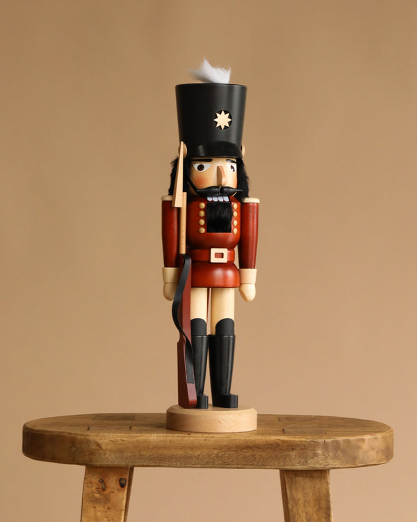 The Christian Ulbricht Collectible Wooden Nutcracker - Soldier, in a red uniform and black hat, stands on a wooden stool against a beige background.