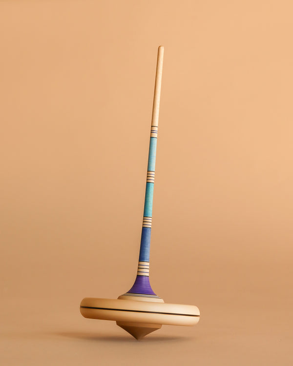 The Mader Arabella Spinning Top - Purple features blue, turquoise, and purple stripes with a long handle and round base. It stands upright on a beige surface and meets strict toy safety standards.
