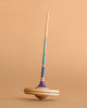 The Mader Arabella Spinning Top - Purple features blue, turquoise, and purple stripes with a long handle and round base. It stands upright on a beige surface and meets strict toy safety standards.