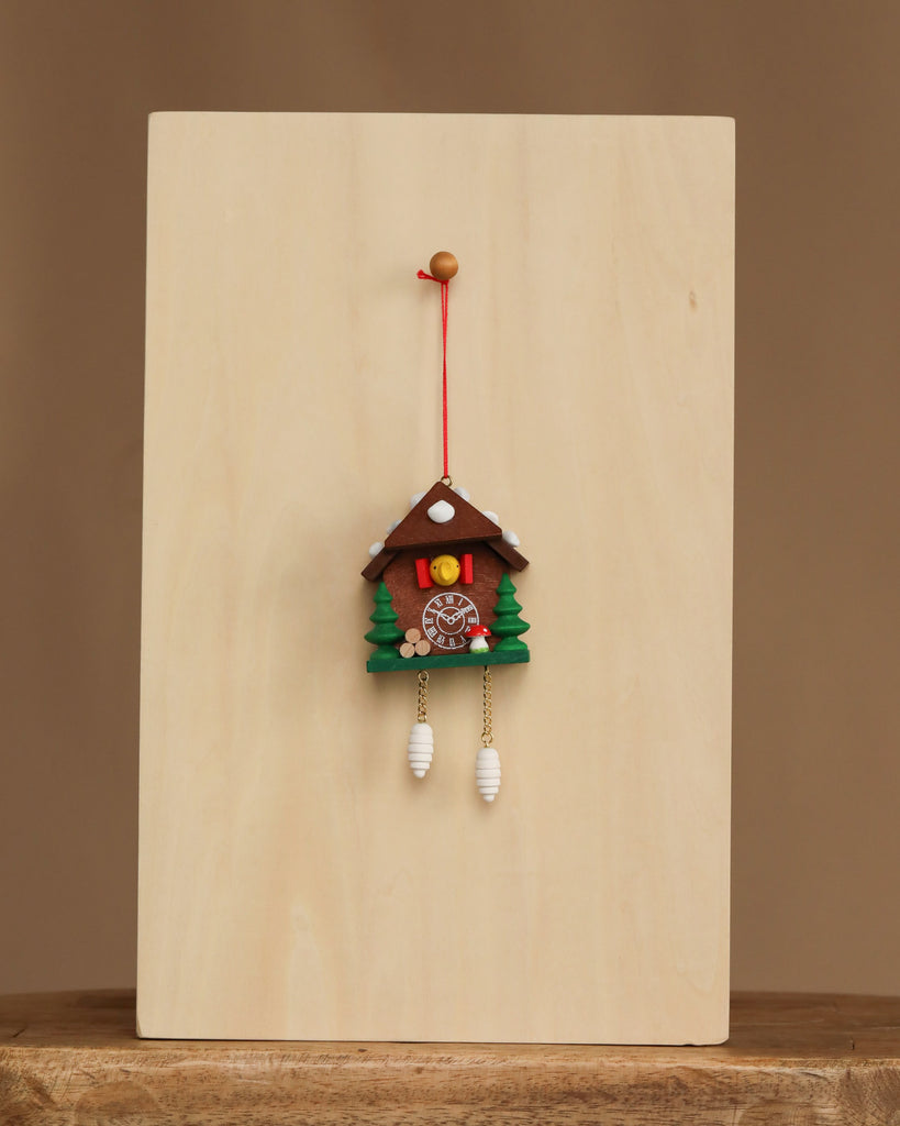 The Christian Ulbricht Ornament - Cuckoo Clock features green trees, a round clock face, and white weights. Hanging on a red string from a wooden board with a beige background. Made in Germany. Contains small parts; choking hazard.