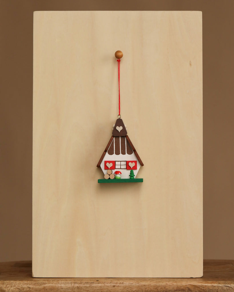The Christian Ulbricht Ornament - Gingerbread House is a small, decorative wooden ornament with heart details, hanging on a red string from a peg on a light wooden board. Contains small parts; choking hazard. Made in Germany.