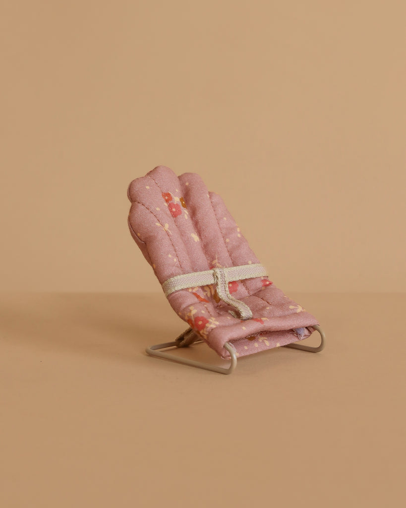 A small, pink padded chair with a white strap and metal legs sits on a light brown background. The Maileg Nursery Set accessory features a subtle floral patterned fabric, making it perfect for imaginative play.