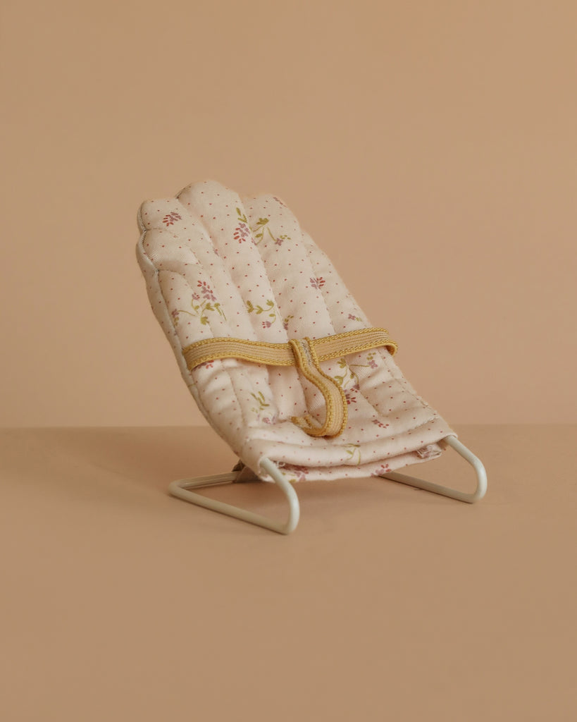 The Maileg Babysitter, Micro - Off White features a small, padded seat with a floral pattern and yellow safety straps on a white metal frame. Perfect as baby doll furniture or for micro-sized rabbits; displayed on a beige surface and background.