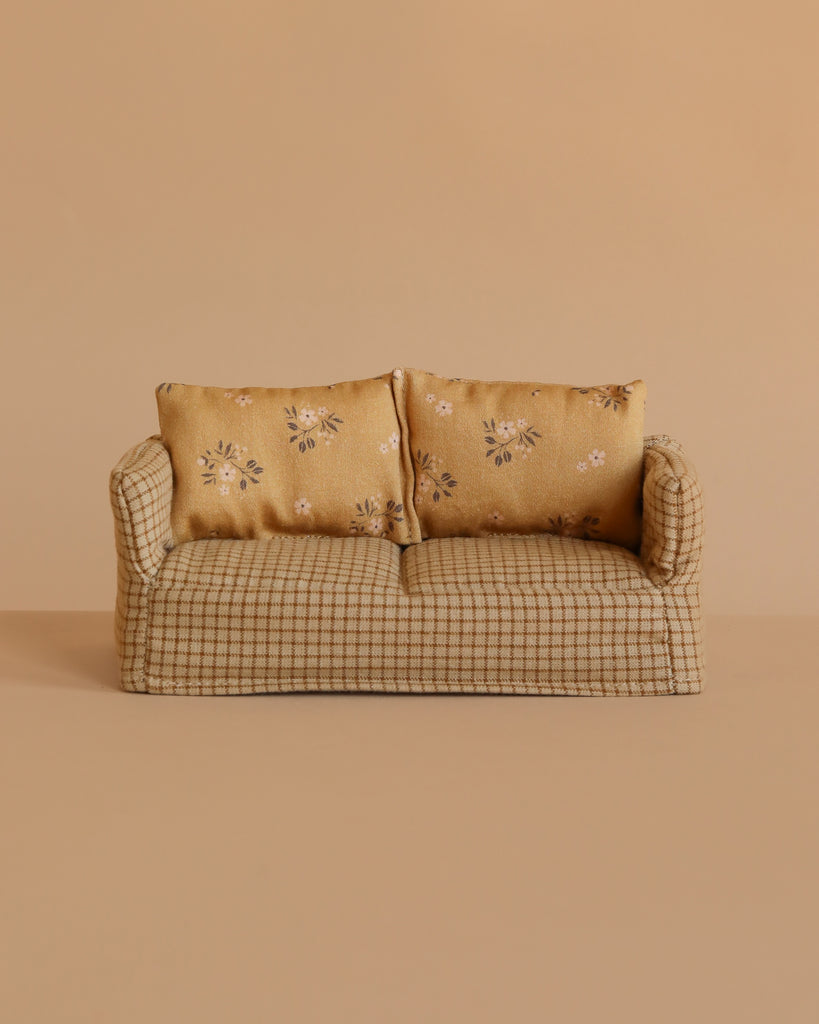 A small beige sofa with plaid fabric and two floral-print cushions, set against a plain tan background, provides an inviting spot for the Maileg Grandparents & Me Set to relax together—perhaps with their cozy cigar box nearby.