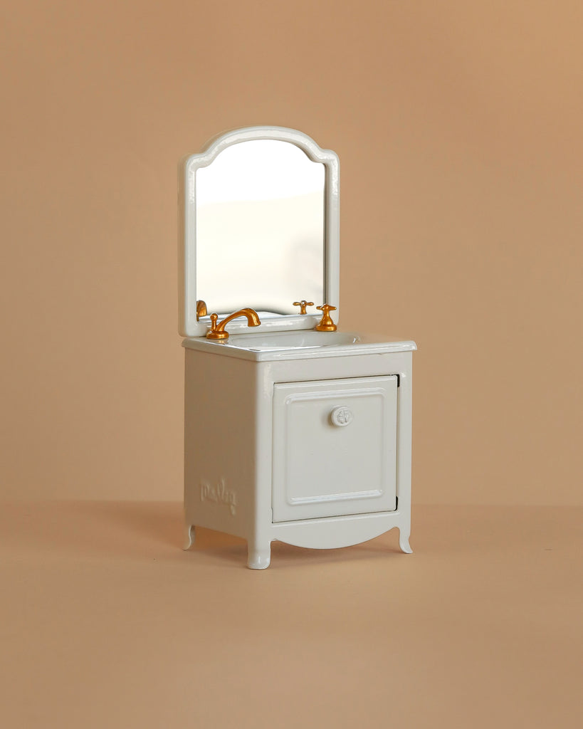 The Maileg Sink Dresser, Mouse Size - Off White, features a vintage look with a mirror and gold faucet handles—ideal for finishing your Mouse hole Farmhouse bathroom scene. Shown beside a small sink on a plain beige background.