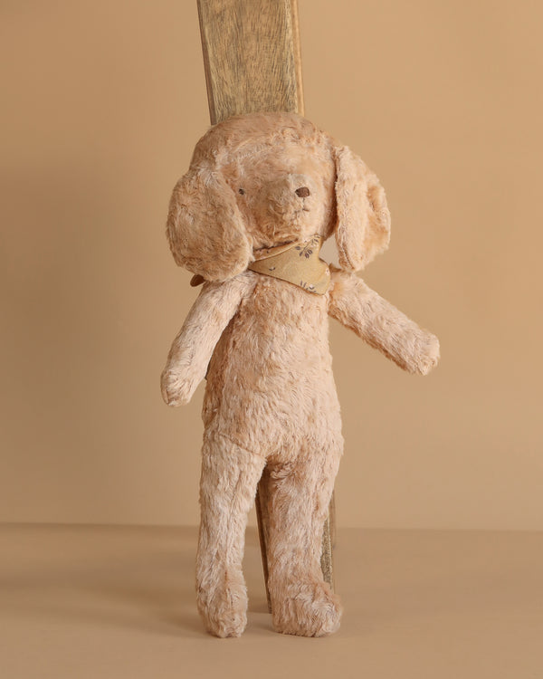 The Maileg Poodle Dog, Vintage Plush in Dusty Powder, a light brown stuffed dog with floppy ears and a scarf, leans against a wooden plank, set against a plain beige background.