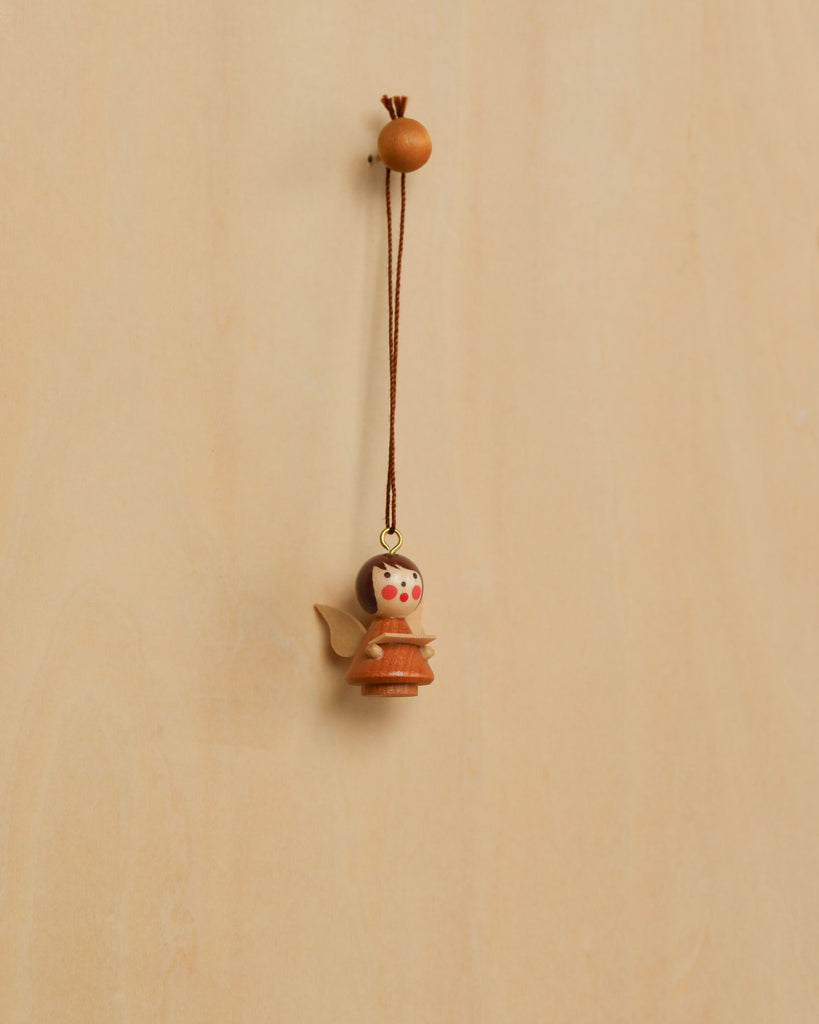 The Christian Ulbricht Ornament - Angel (Natural) is a small wooden angel with painted red cheeks, hanging from a brown string. Made in Germany, this decoration contains small parts and may pose a choking hazard.