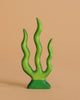 The Handmade Holzwald Seaweed, a sustainable wooden toy painted green with four wavy branches, stands upright against a plain tan background.