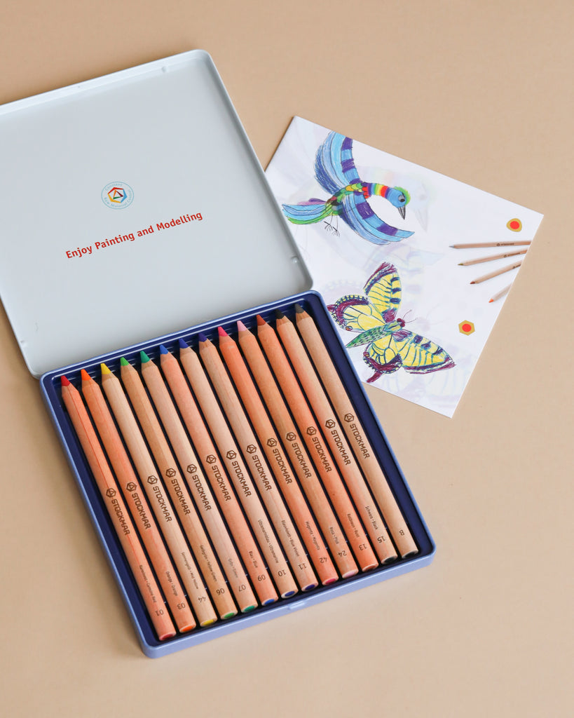 The Moulin Roty Botanist Coloring Book with Stockmar Pencil Set is open next to an illustrated card featuring colorful birds, with a few colored pencils resting on it—all arranged on a beige surface.