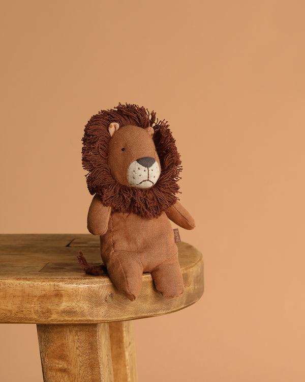The Maileg Lion, Mini—made from soft fabric with a fluffy mane—rests on a wooden stool against a light brown background, ready to join your safari friends.