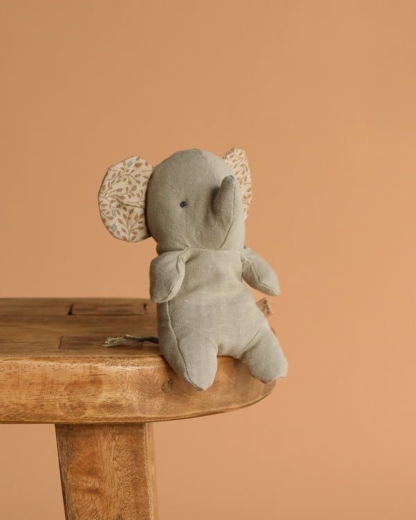 A Maileg Elephant, Mini - Iron Grey plush with patterned ears made from soft fabric sits on the edge of a wooden stool against a plain, light brown background.