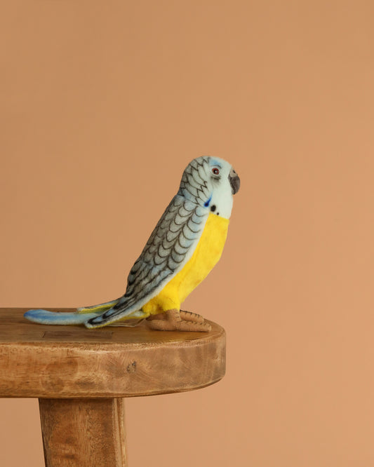 The Parakeet Bird Stuffed Animal, featuring blue, yellow, and black markings, sits lifelike on a wooden table against a beige background, highlighting the realistic detail typical of HANSA animals.