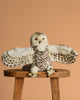 The Snowy Owl Puppet, a hand-sewn plush toy with realistic white and brown patterned feathers and spread wings, rests on a wooden stool against a plain light brown background.