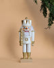 A Christian Ulbricht Collectible Wooden Nutcracker - King (White) stands upright against a beige background. It holds a gold staff and features a red and white checkered bow tie, while pine branches frame the top right corner.