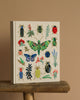 The Curio Embroidered Fabric Sketchbook features a cream hardcover adorned with vibrant embroidered insects and plants, displayed upright on a rustic wooden surface against a plain beige background.
