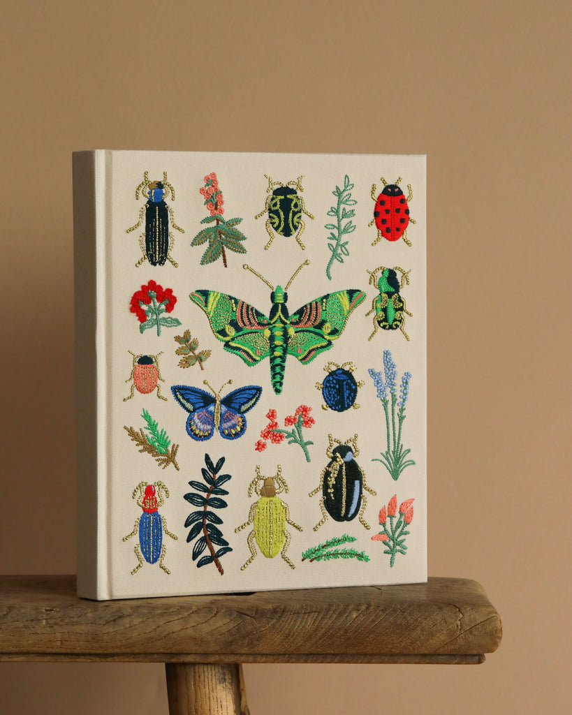 The Curio Embroidered Fabric Sketchbook features a cream hardcover adorned with vibrant embroidered insects and plants, displayed upright on a rustic wooden surface against a plain beige background.