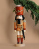The Christian Ulbricht Collectible Wooden Nutcracker - Soldier stands upright, dressed in a red and gold uniform with a tall hat embellished with a white plume and gold trim. Its sword is positioned at its side, while a green pine branch above provides contrast against the beige background.