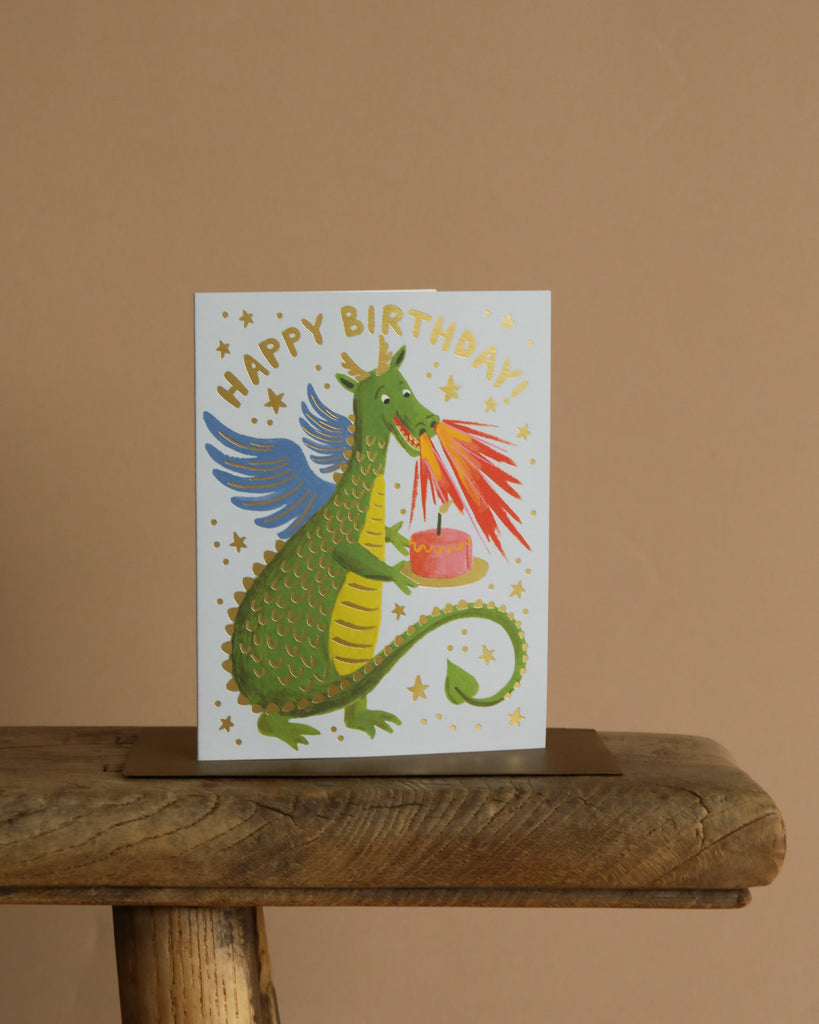 The Birthday Dragon Card features a green winged dragon breathing fire onto a pink cake, surrounded by gold foil stars and "HAPPY BIRTHDAY!" text. Presented on a wooden surface with a beige background. Blank inside.