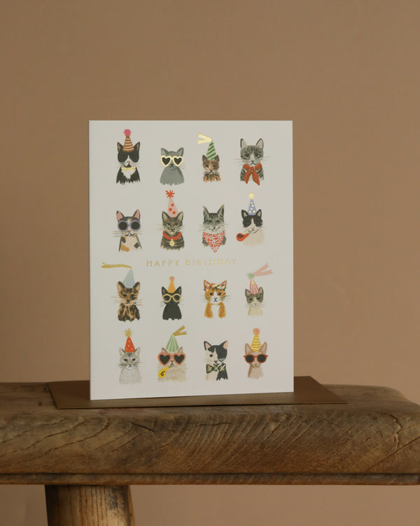 The Cool Cats Birthday Card features illustrated cats in party hats, glasses, and bow ties with metallic gold foil accents and "Happy Birthday" on the cover. The card has a blank interior for your message. Shown on a wooden surface against beige.