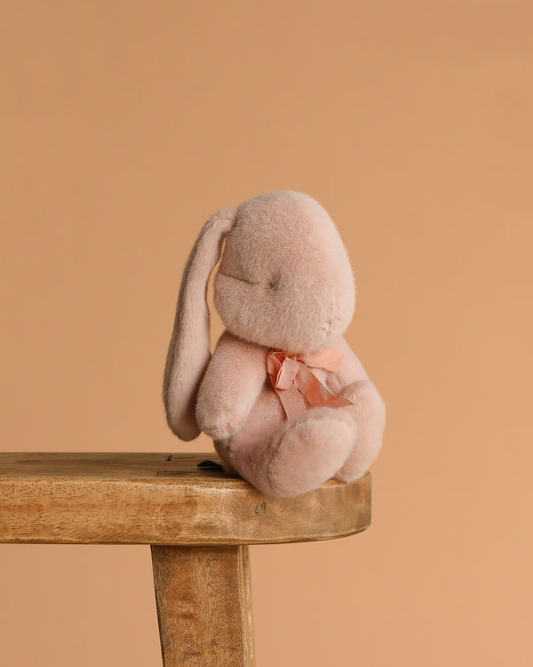 The Maileg Plush Bunny, Small - Powder, with a light pink hue and a bow around its neck, rests on the edge of a wooden stool against a plain peach background, its soft fabric inviting gentle hugs.