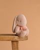 The Maileg Plush Bunny, Small - Powder, with a light pink hue and a bow around its neck, rests on the edge of a wooden stool against a plain peach background, its soft fabric inviting gentle hugs.