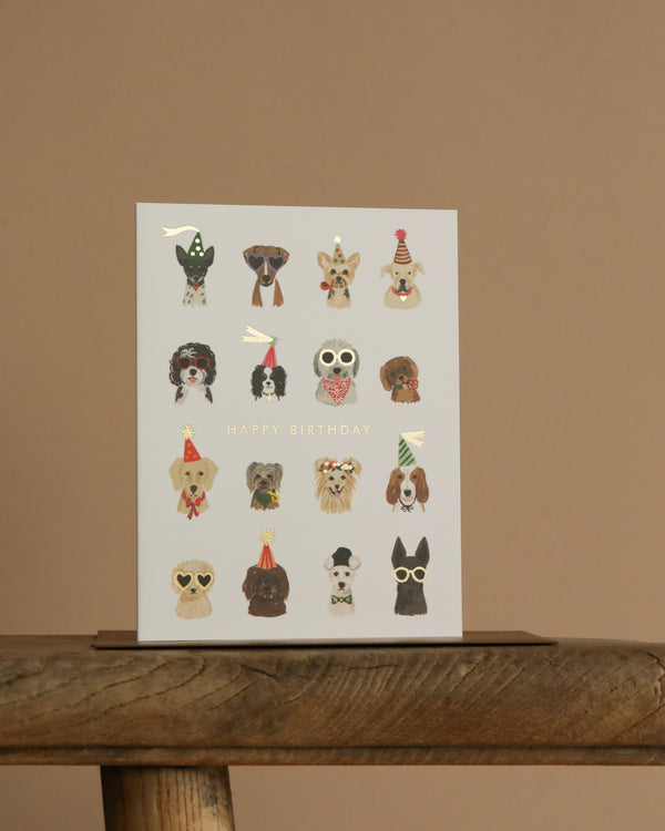 The Party Pups Birthday Card features illustrated dogs in party hats and accessories, arranged in four rows on a plain background. "Happy Birthday" is printed in metallic gold foil at the center, with a blank interior for your message.