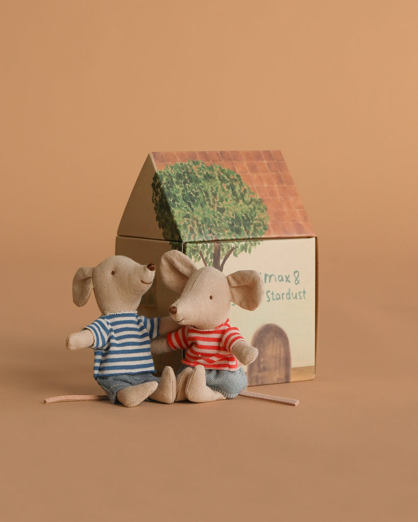 Two plush mice from the Maileg Road Trip Adventure Set sit side by side in striped shirts before a small cardboard dolls house, illustrated with a hand-drawn tree and door, all set against a plain light brown background.