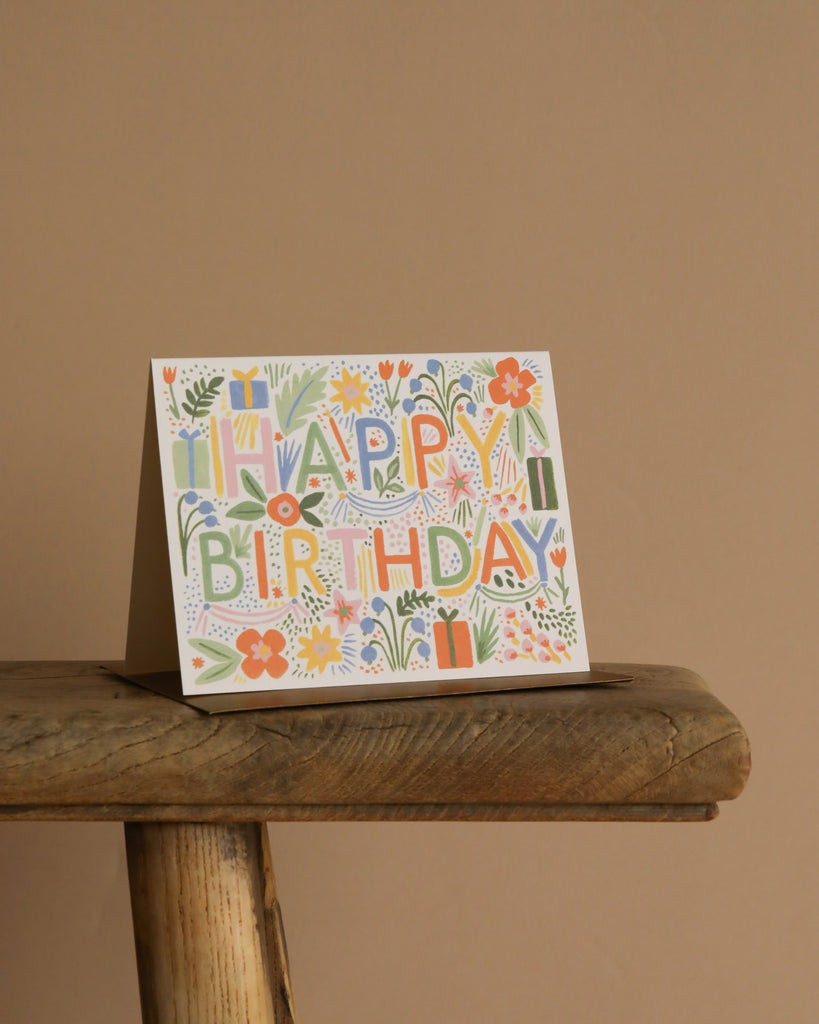 The Fiesta Birthday Card, featuring vibrant floral and gift artwork, stands on a rustic wooden stool with a metallic gold envelope against a plain beige background.