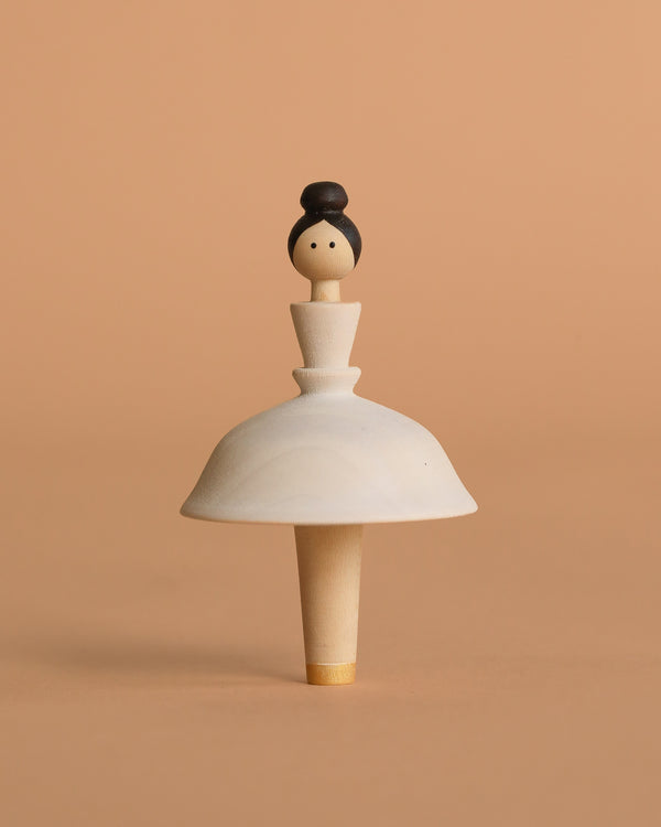 The Sabo Concept Wooden Ballerina is a handmade spinning top crafted to look like a woman with a bun and a white skirt, set against a beige background.