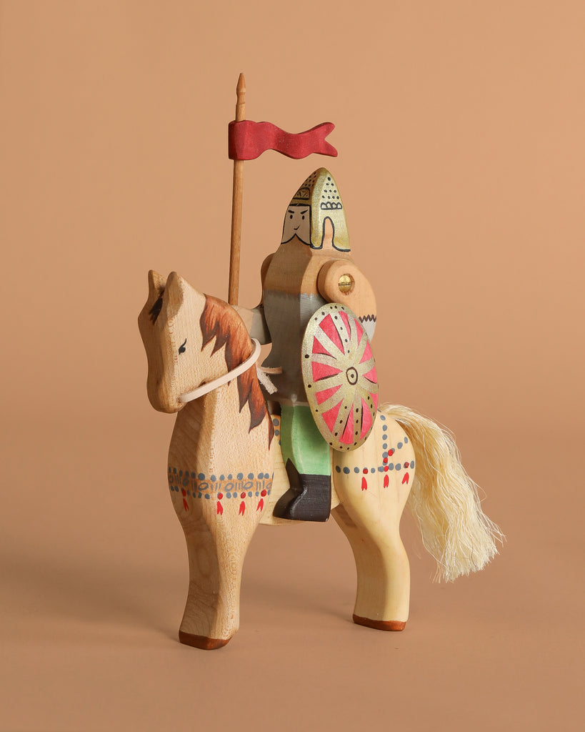 The Bumbu Handmade Wooden Dacian Knight and Steed Set features a painted wooden knight with armor, helmet, red flag, and shield on a yarn-tailed horse—a charming historical toy displayed against a plain beige background.