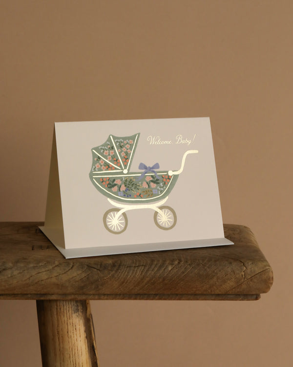 The Floral Pram Card features an illustrated baby stroller with floral patterns, a bow, gold foil accents, and "Welcome Baby!" on the front. It comes with a soft white envelope and is perfect for celebrating new arrivals.