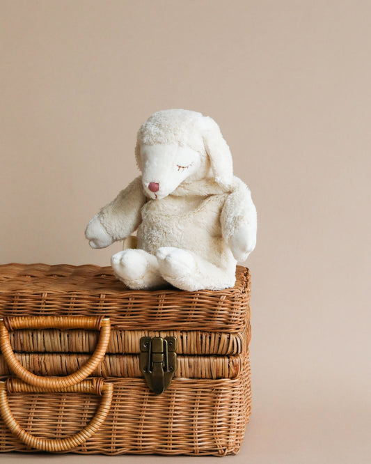 A Senger Naturwelt Cuddly Animal - White Sheep with floppy ears and closed eyes sits on a wicker picnic basket against a soft beige background.