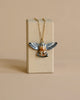 Kestrel Falcon Necklace featuring a gold chain and a handcrafted porcelain falcon pendant with outstretched wings painted in black and white, elegantly displayed on a rectangular beige stand against a beige background—an exquisite piece of limited-edition jewelry.