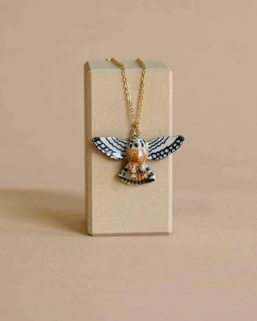 Kestrel Falcon Necklace featuring a gold chain and a handcrafted porcelain falcon pendant with outstretched wings painted in black and white, elegantly displayed on a rectangular beige stand against a beige background—an exquisite piece of limited-edition jewelry.