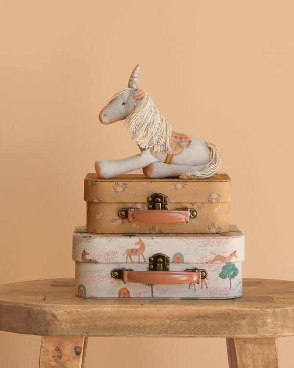 The Maileg Unicorn with Suitcase Set features a plush unicorn sitting on two floral and animal-print suitcases, all displayed on a wooden table with a peach-colored background.