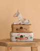 The Maileg Unicorn with Suitcase Set features a plush unicorn sitting on two floral and animal-print suitcases, all displayed on a wooden table with a peach-colored background.