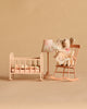 Two stuffed mice sit on a peach rocking chair next to a lamp and an empty Maileg Cradle, My - Light Rose—complete with a blanket and pillows—all set against a beige background.