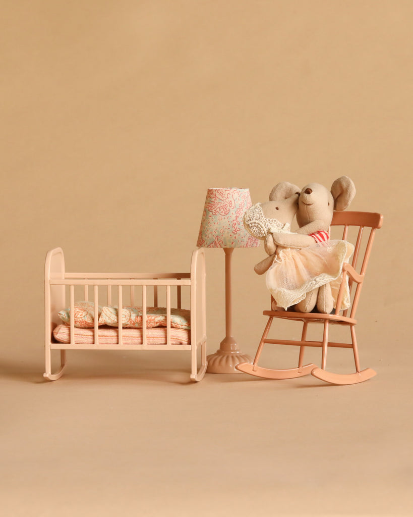 Two stuffed mice sit on a peach rocking chair next to a lamp and an empty Maileg Cradle, My - Light Rose—complete with a blanket and pillows—all set against a beige background.