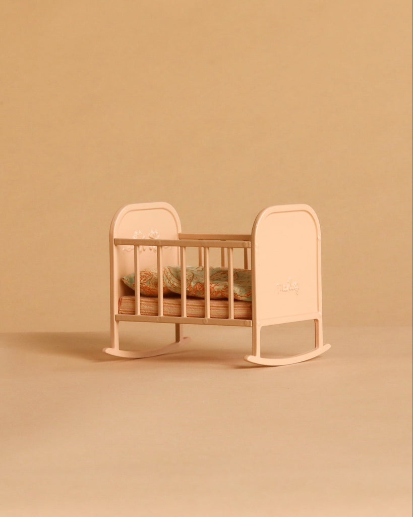 The Maileg Cradle, My - Light Rose, featuring floral bedding, sits atop a beige surface with a matching beige background.