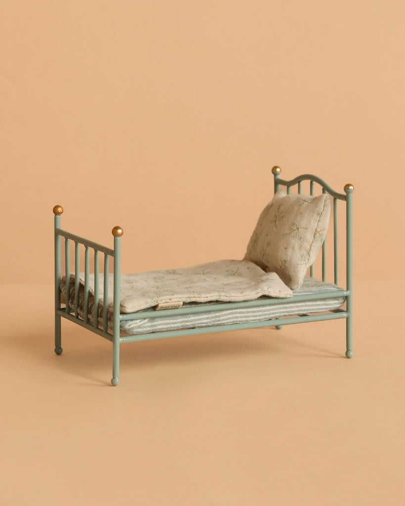 The Maileg Little Lambs Bedroom Set features a mint-green vintage single bed with gold-tipped posts, a light gray floral pillow, and a matching blanket—ideal for any Maileg Dollhouse.