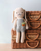 Cuddle + Kind Henry the Bunny is a hand-knit organic cotton doll in grey overalls with a carrot design, leaning against wicker baskets—an adorable, gentle-play toy.