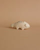 A small, hand-carved Albizia wood handmade tiny wooden animals - chameleon figurine, crafted by skilled artisans in Bali, features simple black line details for eyes, a mouth, and patterned markings on its back. Set against a peach-colored background, the light beige chameleon has a charming curved tail.