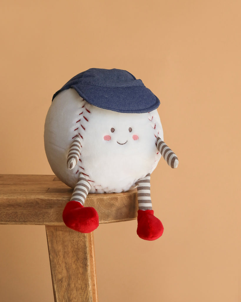 The Mon Ami - Buddy Baseball Plush Toy, featuring a smiling face, striped arms and legs, red shoes, and a blue cap, sits on the edge of a wooden bench against a beige background.