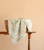An Organic Cotton Baby Blanket - Fishing is draped over a wicker basket that sits atop a wooden table against a plain peach background. The light-colored, patterned blanket—crafted in Portugal—introduces a touch of gentle elegance to the scene.