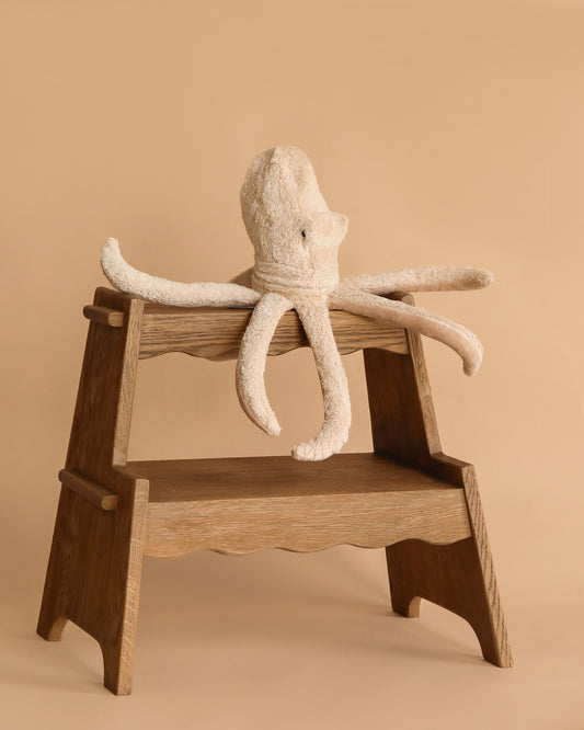 The Senger Naturwelt Cuddly Animal - Octopus, made from organic cotton with long floppy arms, rests on a wooden step stool against a beige background—an ideal calming companion for cozy moments.